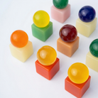 Levendige, doorschijnende bollen rusten op effen pastelkleurige kubussen, keurig gerangschikt in een raster op een witte achtergrond. Elke bol heeft een unieke kleur en rust op een bijpassende of contrasterende kubus, wat doet denken aan het speelse ontwerp en de levendige tinten van de Matias Moellenbach OBA Zeep Bar Papaya van Matias Moellenbach.