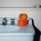Een blauwe wasbak met waterdruppels, verchroomde kranen, een oranje stuk zeep met daarop een doorzichtige oranje bol, en twee ronde beige bakjes op een witte plank op de achtergrond vormen een decor dat het sculpturale ontwerp benadrukt. De opvallende Matias Moellenbach OBA Zeep + Houder Papaya van Matias Moellenbach vult de ruimte met een subtiele papajageur voor een verfrissend tintje.