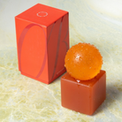 Een levendig oranje bol, die doet denken aan de Matias Moellenbach OBA Zeep + Houder Papaya, glinstert met waterdruppeltjes bovenop de bijpassende geometrische houder en een bijpassende rechthoekige doos, allemaal geplaatst op een licht marmeren oppervlak — waarmee de sculpturale stijl van Matias Moellenbach wordt weergegeven en een verfrissende vleug papaja wordt opgeroepen.