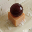 Een ronde, donkerpaarse vrucht ligt op een bubbelachtige, vierkante Matias Moellenbach OBA Zeep + Houder Terralis op een nat badkameroppervlak, met witte tegels op de achtergrond. Dit benadrukt het elegante Scandinavische ontwerp van Matias Moellenbach en roept de frisse Terralis-geur op.