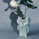 Een witte roos met groene bladeren zit in de Serax After Midnight Uil Vaas Lichtblauw S, een lichtblauwe keramische vaas in de vorm van een uil van Serax, tegen een grijs gestructureerde achtergrond.