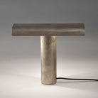 De Serax Fck Tafellamp Staal Grijs van Serax heeft een modern, minimalistisch ontwerp met een cilindrische voet, platte rechthoekige bovenkant, afwerking met metallic textuur en zichtbaar zwart netsnoer op een wit oppervlak.