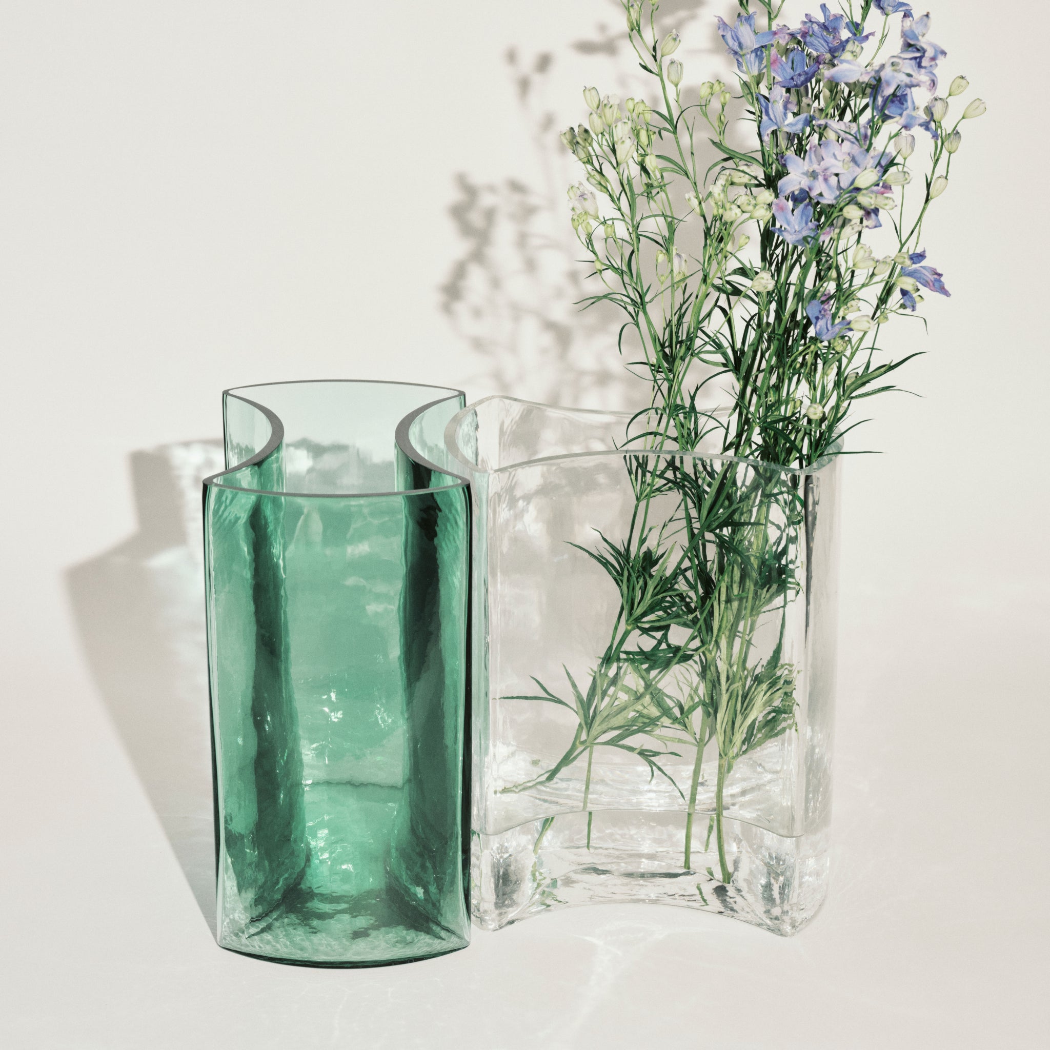 Twee glazen vazen - een Serax Parole Vaas Helder en een groene vaas - staan naast elkaar. De heldere Serax vaas toont een boeket paarse en witte wilde bloemen met groen, die zachte schaduwen werpen op een lichte achtergrond.