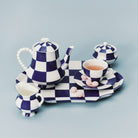 Het Serax Wouters & Hendrix Fête Des Bêtes Melkkannetje Blauw Wit wordt getoond als onderdeel van een blauw-wit geblokt Serax theeservies, gerangschikt met roze snoepjes op het schoteltje tegen een lichtblauwe achtergrond.