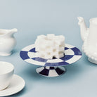 Een stapel suikerklontjes is gerangschikt in een kubusvorm op de Serax Wouters & Hendrix Fête Des Bêtes Taartschaal Blauw Wit, met een witte theepot, creamer en kop vlakbij tegen een lichtblauwe achtergrond.