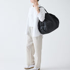 Een persoon in een wit shirt, lichte broek en witte sneakers staat tegen een effen achtergrond met een Shupatto Opvouwbare Boodschappentas Jet Zwart L van Shupatto over zijn schouder. Hun gezicht is gedeeltelijk buiten beeld.