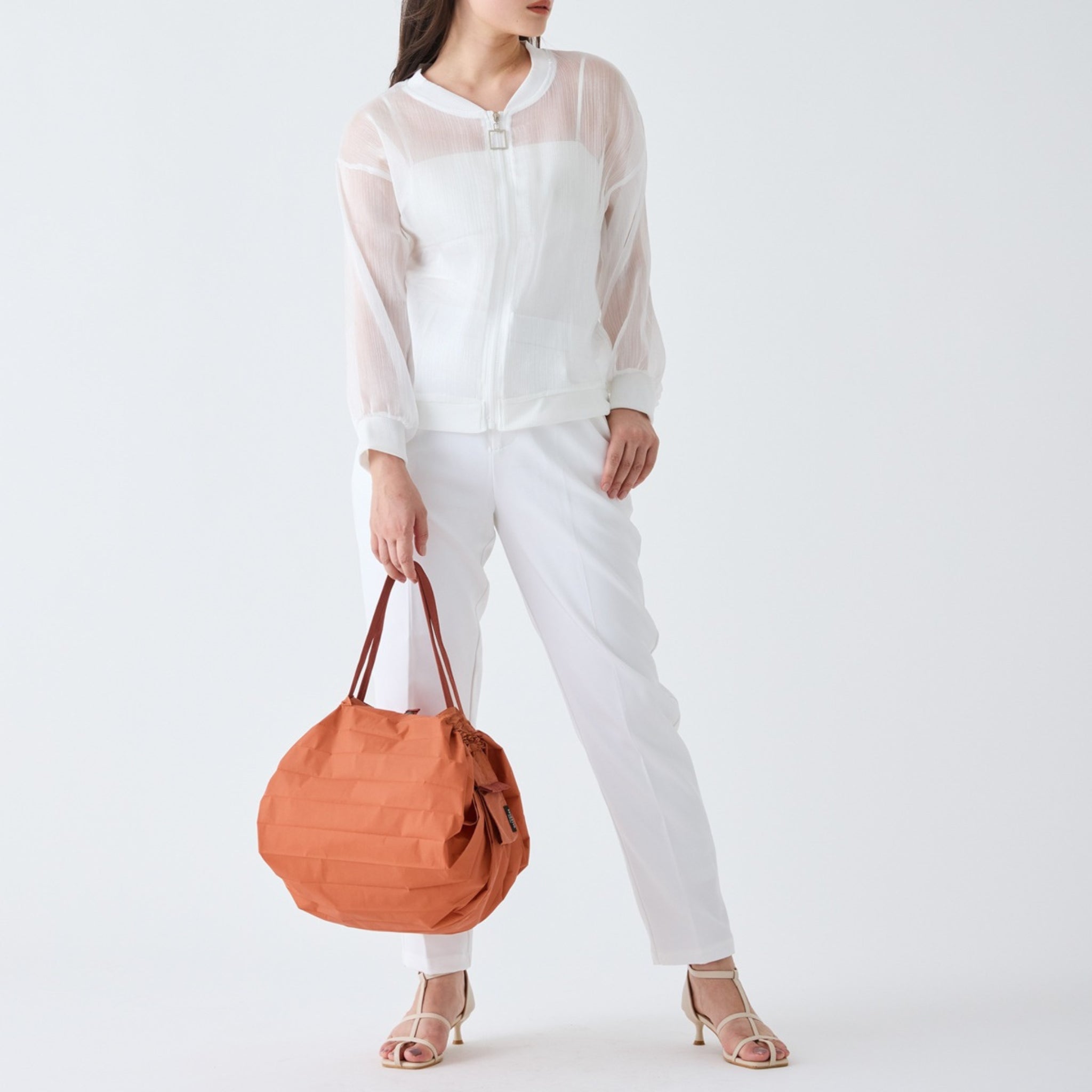 Een persoon in een doorschijnend wit jasje, witte broek en sandalen met hak houdt een grote Shupatto Opvouwbare Boodschappentas Brick Bruin M van Shupatto vast tegen een effen witte achtergrond.