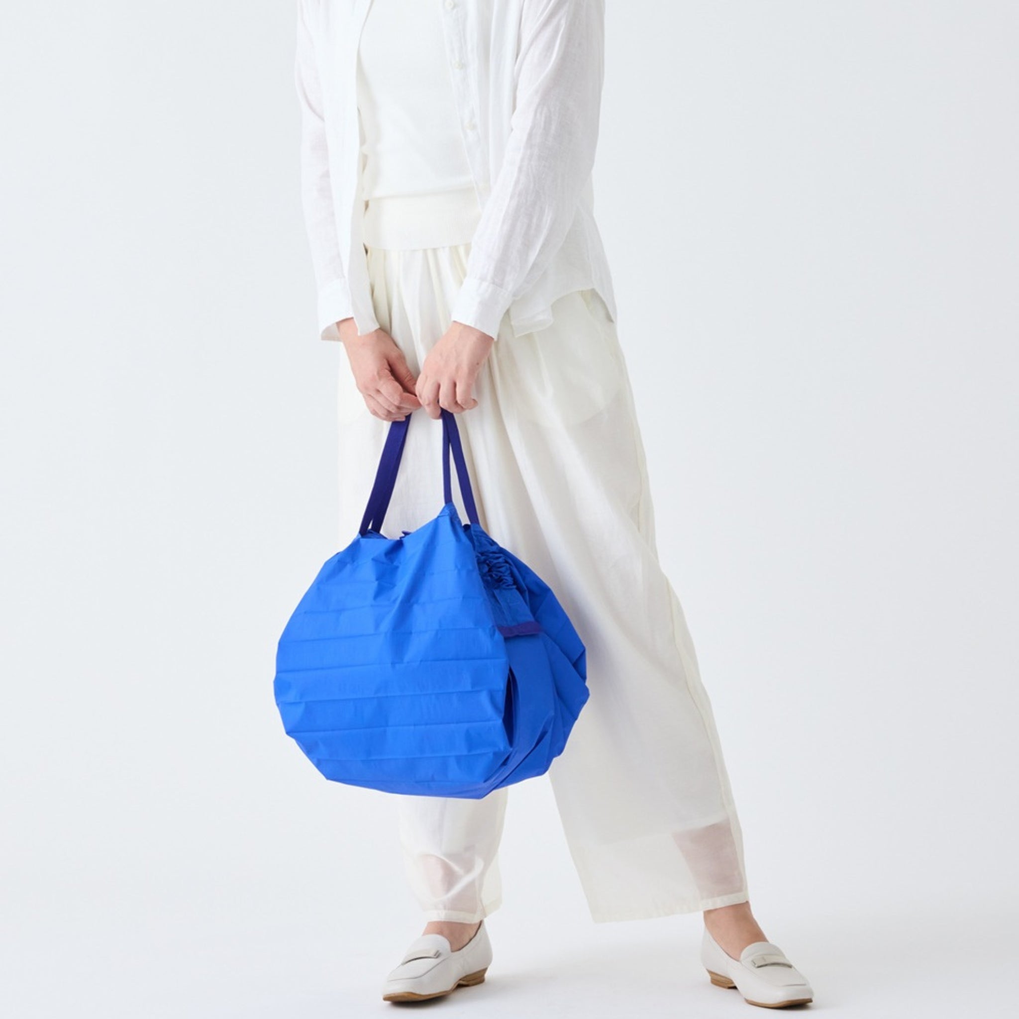 Een persoon in losse witte kleding en schoenen houdt een grote Shupatto Opvouwbare Boodschappentas Clear Sky Blauw M met donkere hengsels van Shupatto vast. De effen witte achtergrond en het onzichtbare gezicht van de persoon benadrukken de tas.