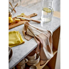 Een Södahl Twist Theedoek Taupe 2 st. en een geel servet rusten op een marmeren aanrecht naast borden, een houten plank met gesneden citroenen en een glazen waterkan - ideaal voor een Scandinavische keukenstijl.
