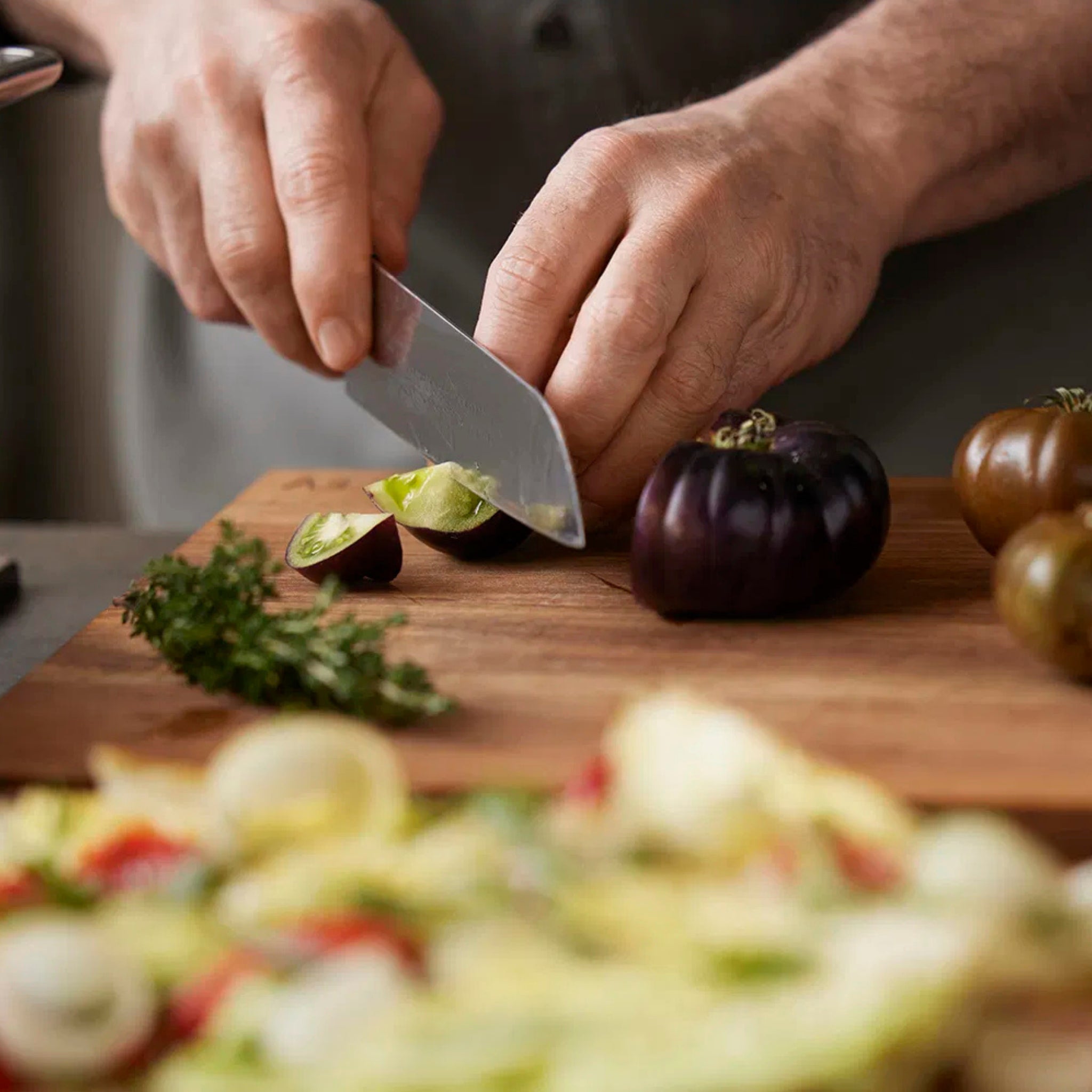 Iemand snijdt een kleine groene tomaat op de Tareq Taylor Tarragon Snijplank Acacia A3 42/30, omringd door verse kruiden en andere tomaten uit de Tarragon collectie, met een vage salade op de voorgrond.