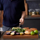 In een moderne keuken geïnspireerd door de Tareq Taylor Dragon collectie, staat een persoon in een marineblauw overhemd achter de Tareq Taylor Tarragon Snijplank Acacia A3 42/30 met gehalveerde paarse aardappelen, koriander, citroenen, limoenen en broodjes.