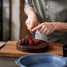Een persoon snijdt een ronde chocoladetaart met daarop verse frambozen, rode bessen en bramen op de Tareq Taylor Tarragon Snijplank Acacia A3 42/30 van Tareq Taylor. Het gezicht van de persoon is niet zichtbaar.