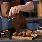 Een persoon met een bruin schort kookt in de keuken en houdt voedsel boven een blauwe pan. Gebakken items rusten op de Tareq Taylor Tarragon Snijplank Acacia A5 21/15, die de kenmerkende gezellige stijl van Tareq Taylor vastlegt.