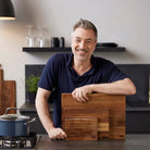 Een lachende man in een marineblauw overhemd staat in een moderne keuken, leunend op het aanrecht terwijl hij drie gestapelde Tareq Taylor Dragon Snijplank Acacia A4 30/21 snijplanken vasthoudt. Vlakbij staat een pan op het fornuis.