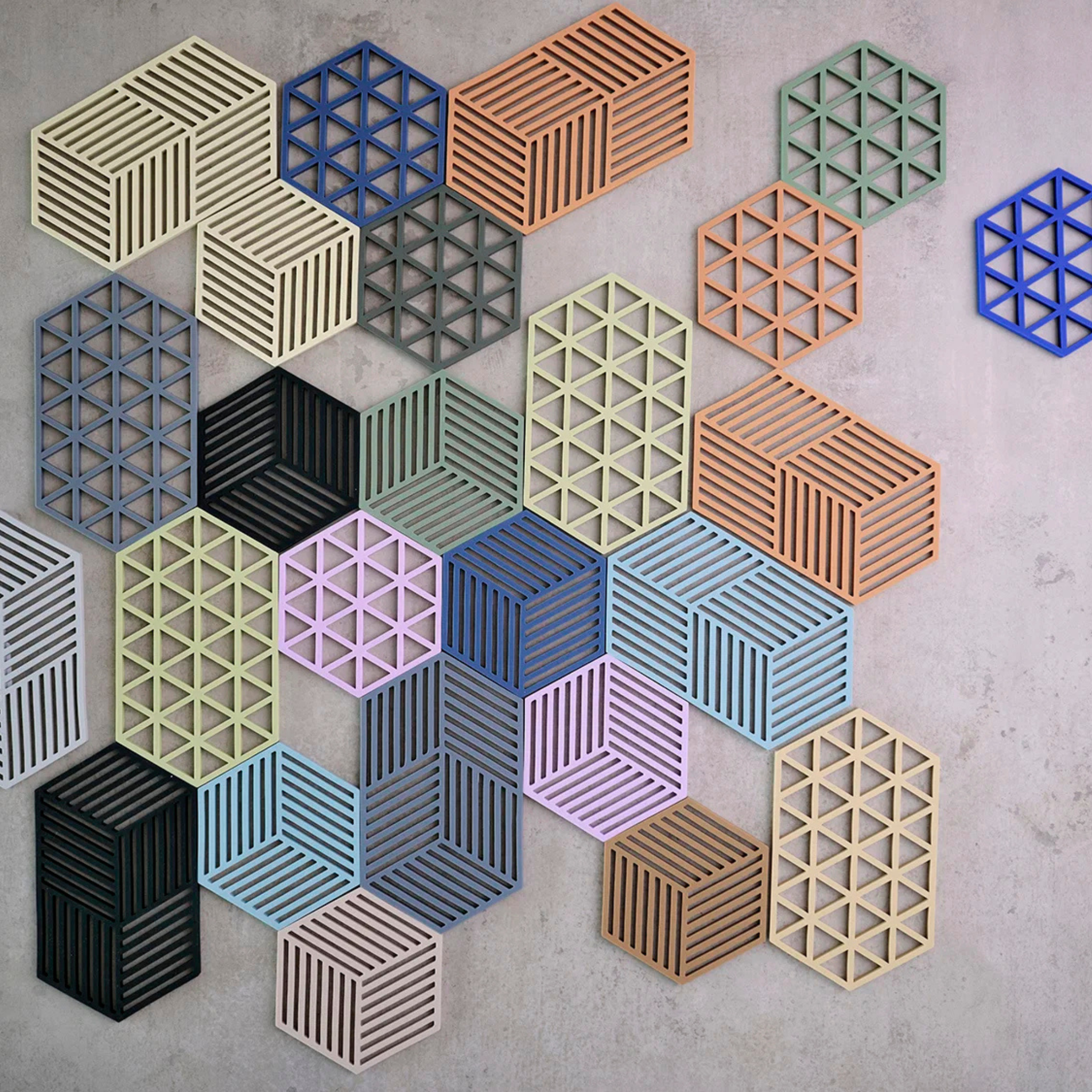 Het Zone Denmark Hexagon Trivet in olijfgroen heeft geometrische pastelkleurige en neutrale zeshoeken en kubussen met gestreepte en driehoekige patronen, die lijken op moderne muurkunst tegen een lichtgrijze betonnen achtergrond.